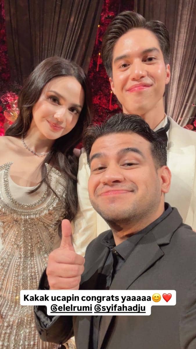 Seorang tamu berpose bersama pasangan pengantin dalam resepsi pernikahan dengan latar dekorasi bunga merah dan pencahayaan hangat.