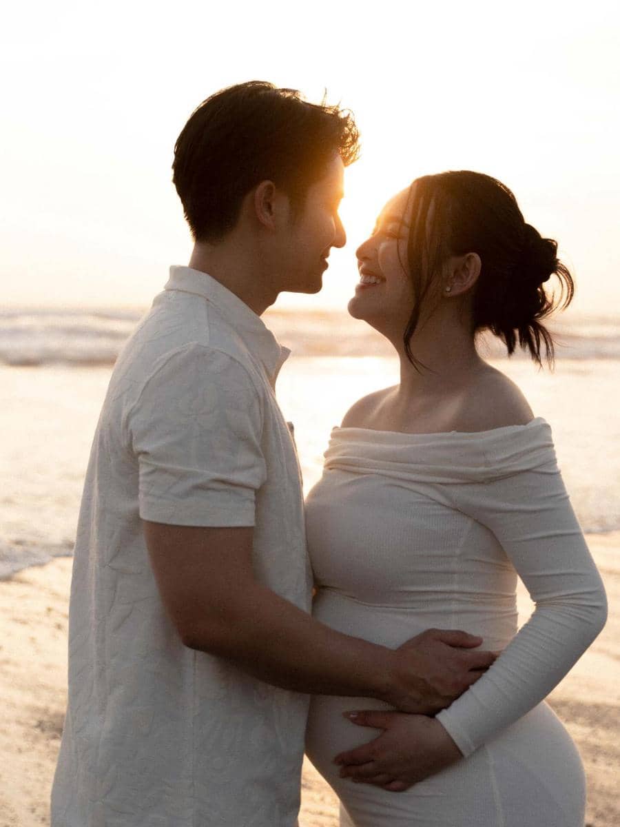 Pasangan berpose mesra di tepi pantai saat matahari terbenam, pria memegang perut wanita hamil yang mengenakan gaun putih.