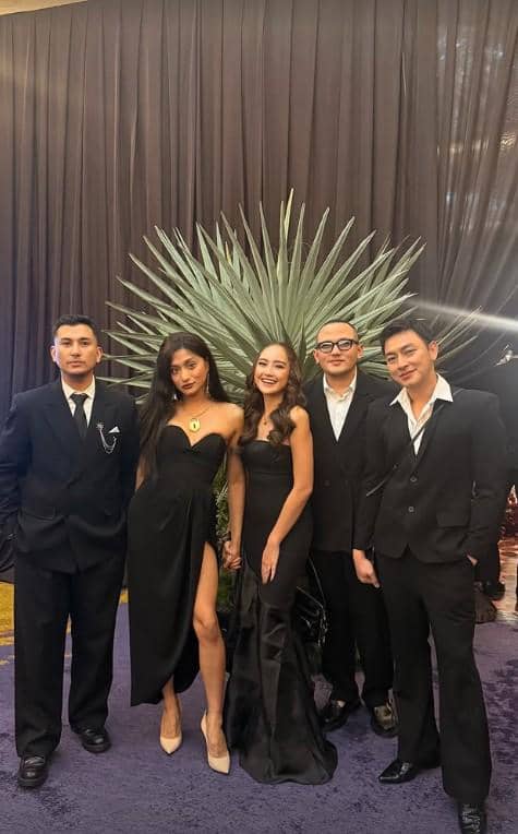 Lima tamu berpakaian formal berpose bersama di acara resepsi pernikahan dengan latar dekorasi tanaman hijau dan tirai hitam elegan.