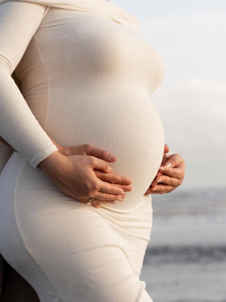 Perempuan hamil mengenakan gaun rajut berwarna krem memegang perutnya dengan lembut di tepi pantai saat pemotretan maternity.