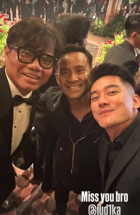 Tiga pria berpakaian formal berpose bersama di acara resepsi pernikahan dengan latar dekorasi bunga dan tamu undangan.