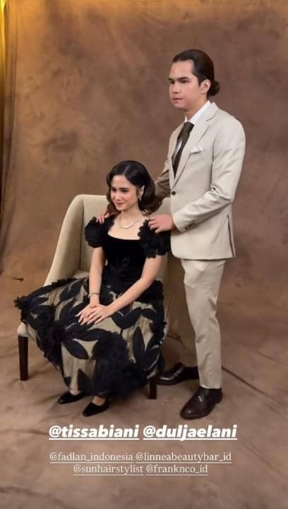 Seorang pria mengenakan setelan jas krem berdiri di samping wanita bergaun hitam dan krem yang duduk di kursi dengan latar cokelat elegan.