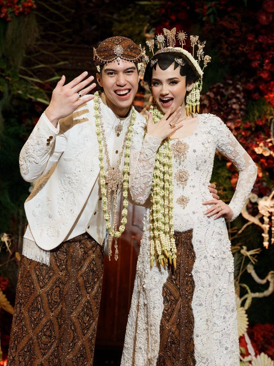 Sepasang pengantin mengenakan busana adat Jawa berwarna putih dan batik cokelat, tersenyum sambil memperlihatkan cincin pernikahan mereka.