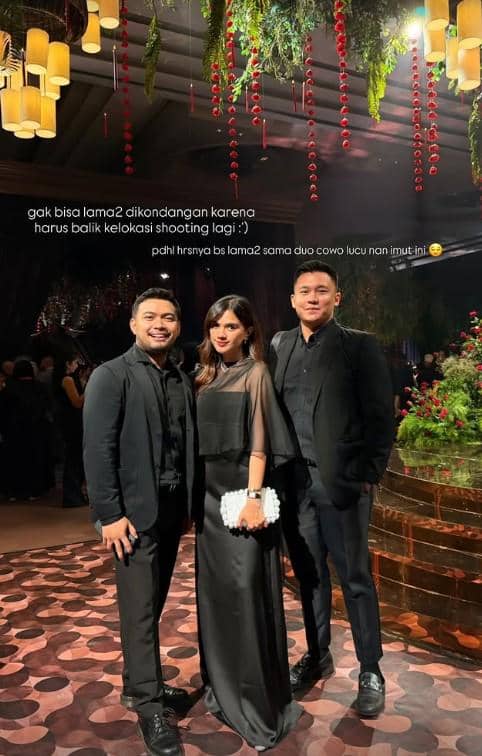 Tiga tamu berpakaian formal hitam berpose di resepsi pernikahan dengan dekorasi bunga gantung dan pencahayaan hangat di ruangan mewah.