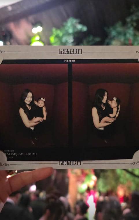 Seorang tamu memegang hasil foto photobooth yang menampilkan seorang wanita bergaun hitam menggendong bayi di resepsi pernikahan.