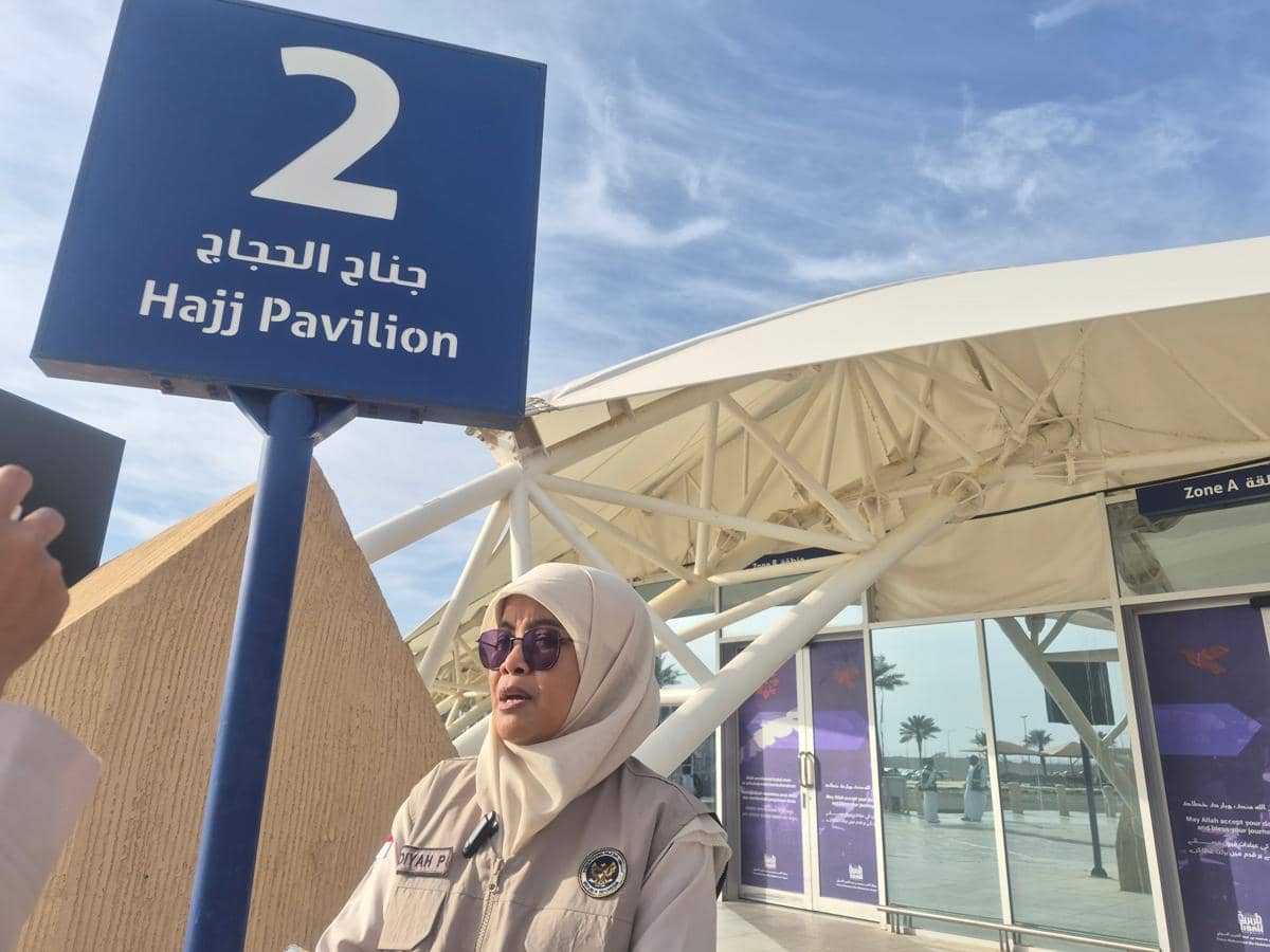 Petugas pembimbing ibadah mengenakan seragam dan hijab krem berdiri di dekat papan bertuliskan Hajj Pavilion 2 di area bandara.