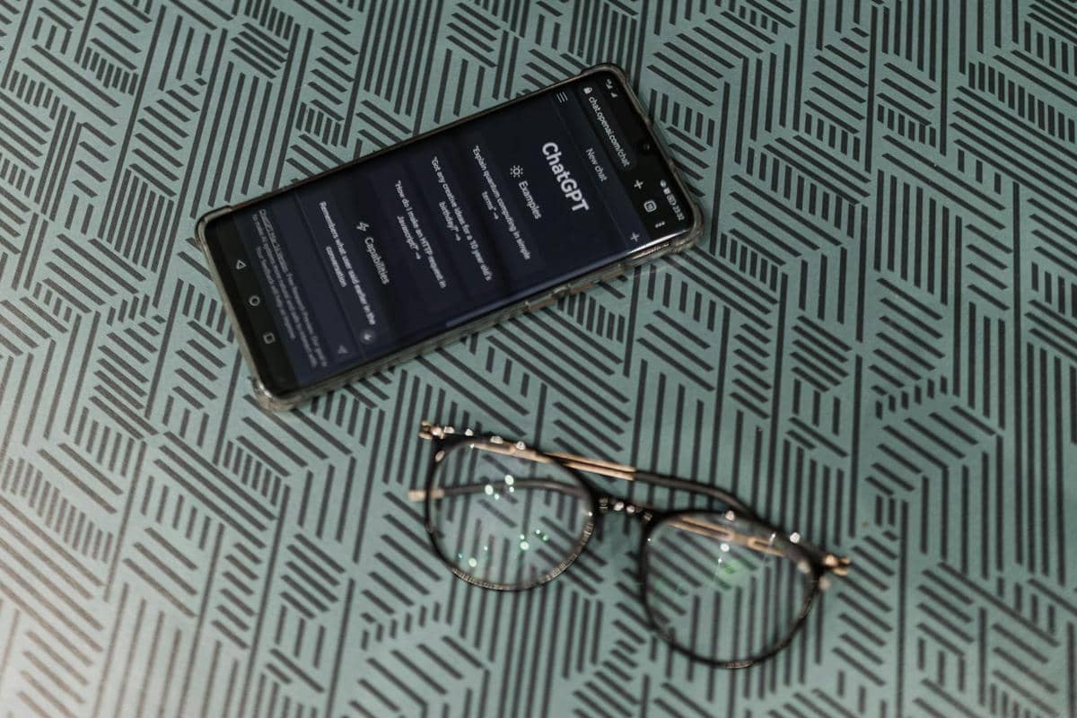 Ponsel menampilkan antarmuka ChatGPT di atas meja bermotif geometris dengan kacamata hitam bulat di sampingnya.