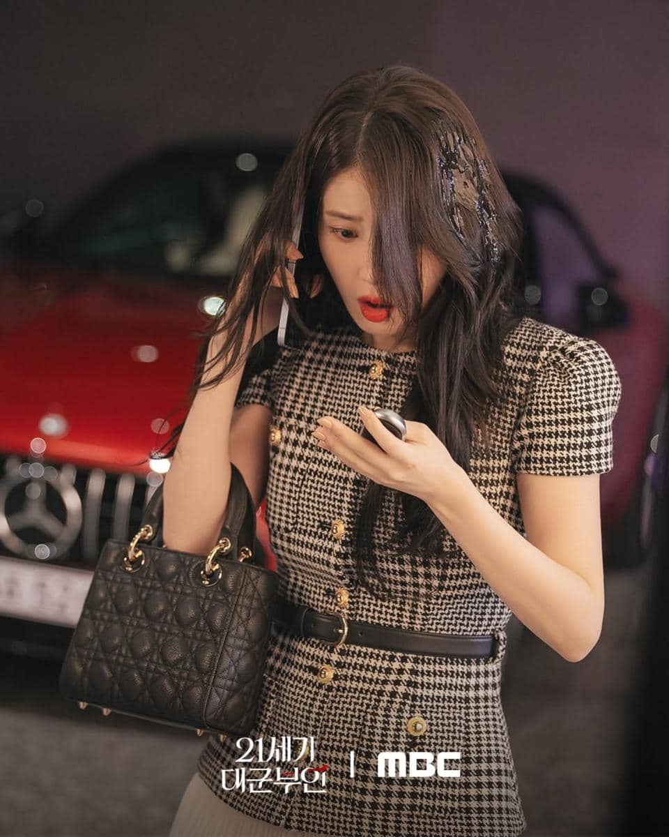 Seorang wanita berpakaian elegan bermotif kotak-kotak memegang ponsel dengan ekspresi terkejut di depan mobil merah.