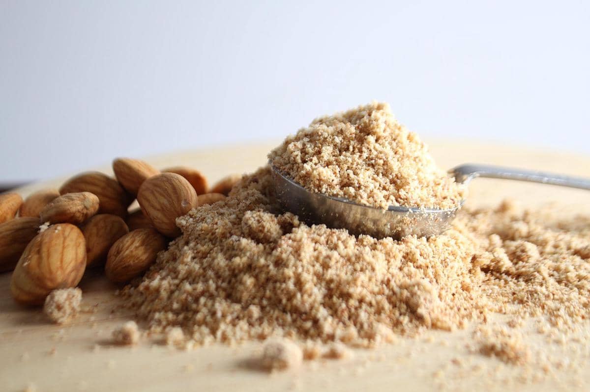Bubuk kacang almond halus di atas meja kayu dengan sendok logam dan beberapa butir almond utuh di sekitarnya.
