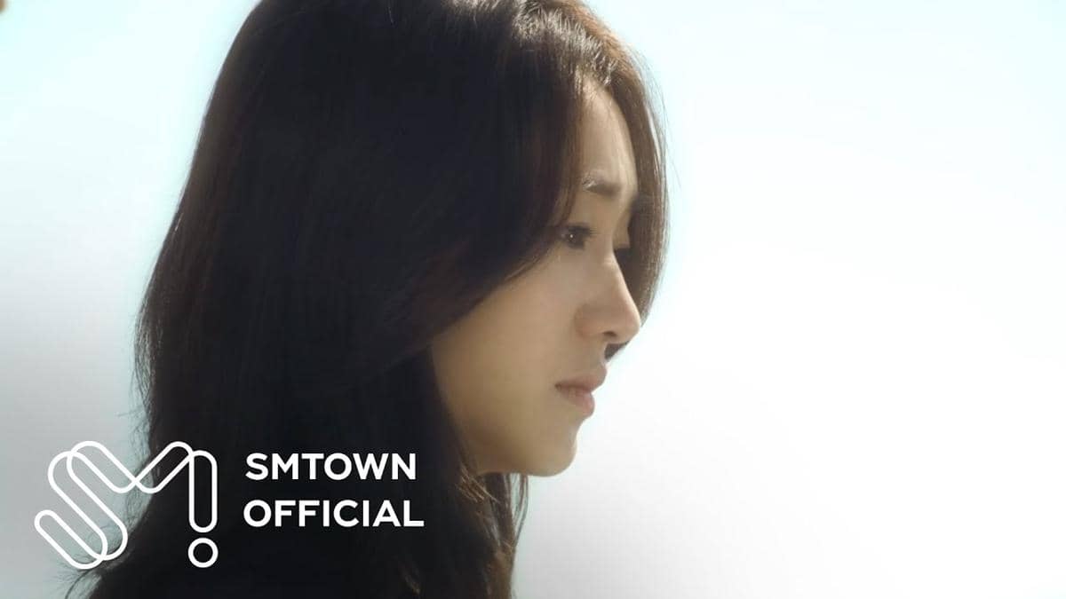 Seorang wanita berambut panjang menatap ke bawah dengan ekspresi sedih di latar belakang langit cerah, dengan logo SMTOWN Official di sudut gambar.