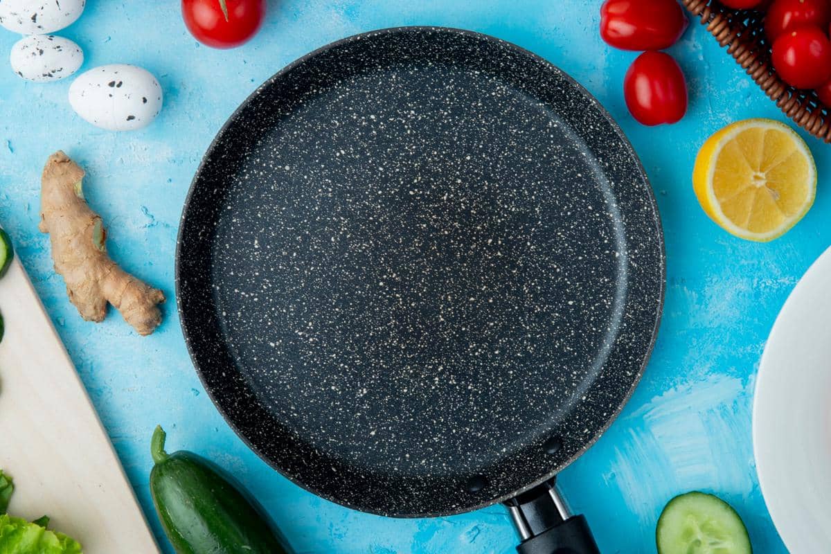 Wajan teflon berwarna hitam dengan motif bintik putih dikelilingi bahan makanan seperti jahe, tomat, lemon, dan telur di atas meja biru.