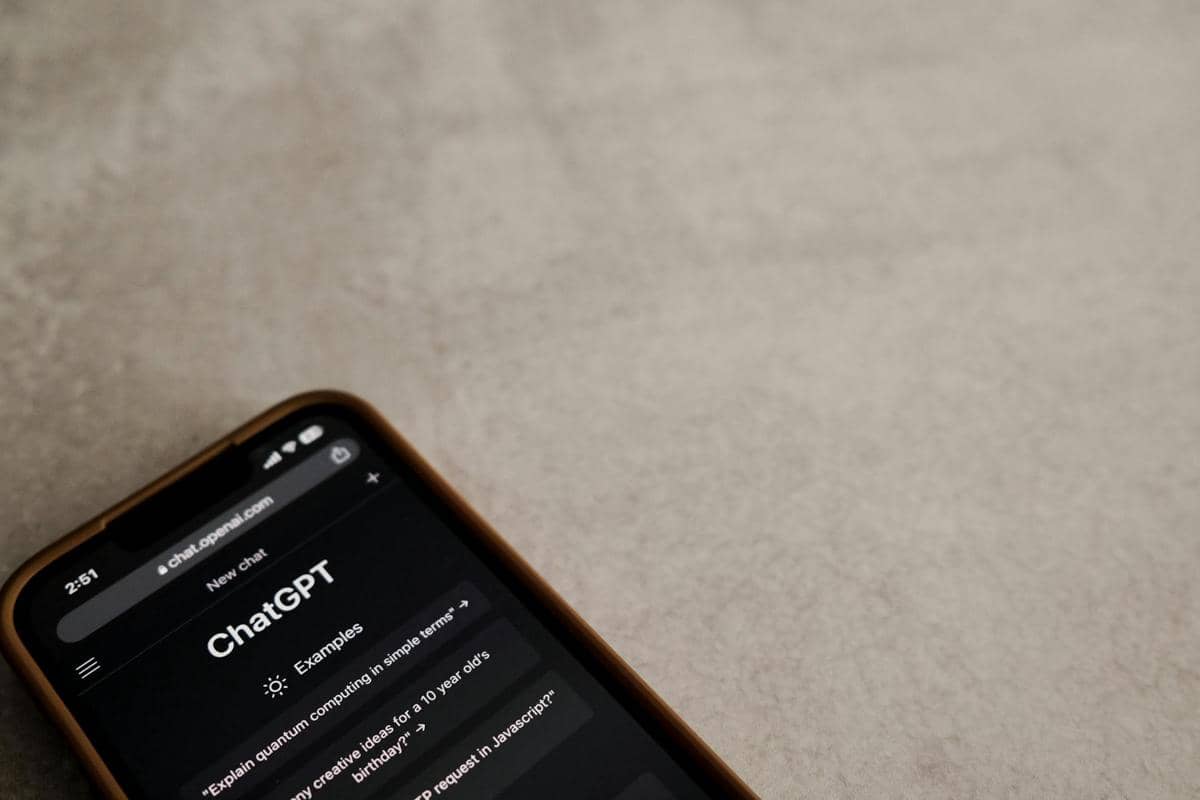 Layar ponsel menampilkan antarmuka ChatGPT di situs chat.openai.com dengan latar permukaan meja berwarna abu-abu.
