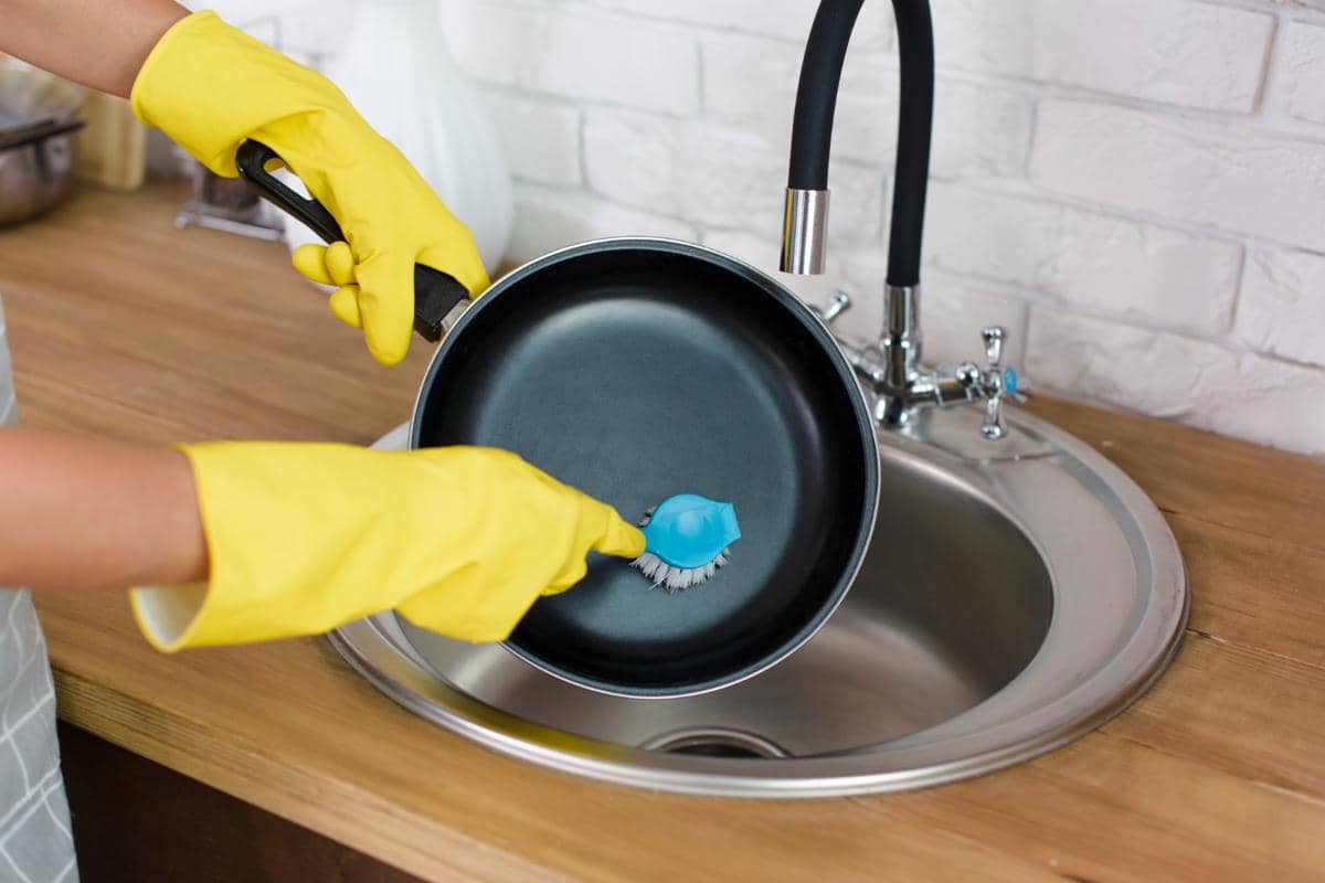 Seseorang mengenakan sarung tangan karet kuning sedang mencuci wajan teflon hitam menggunakan sikat di wastafel dapur.