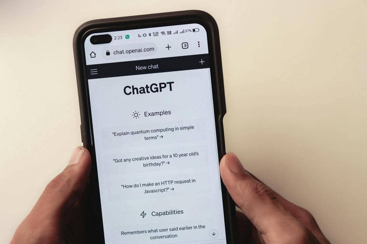Seseorang memegang ponsel yang menampilkan laman ChatGPT di browser dengan antarmuka berwarna putih dan teks contoh percakapan.