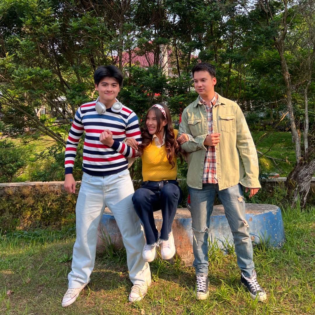 Tiga remaja bergaya kasual berpose di luar ruangan, dua pria mengenakan pakaian santai dan satu wanita mengenakan sweater kuning.