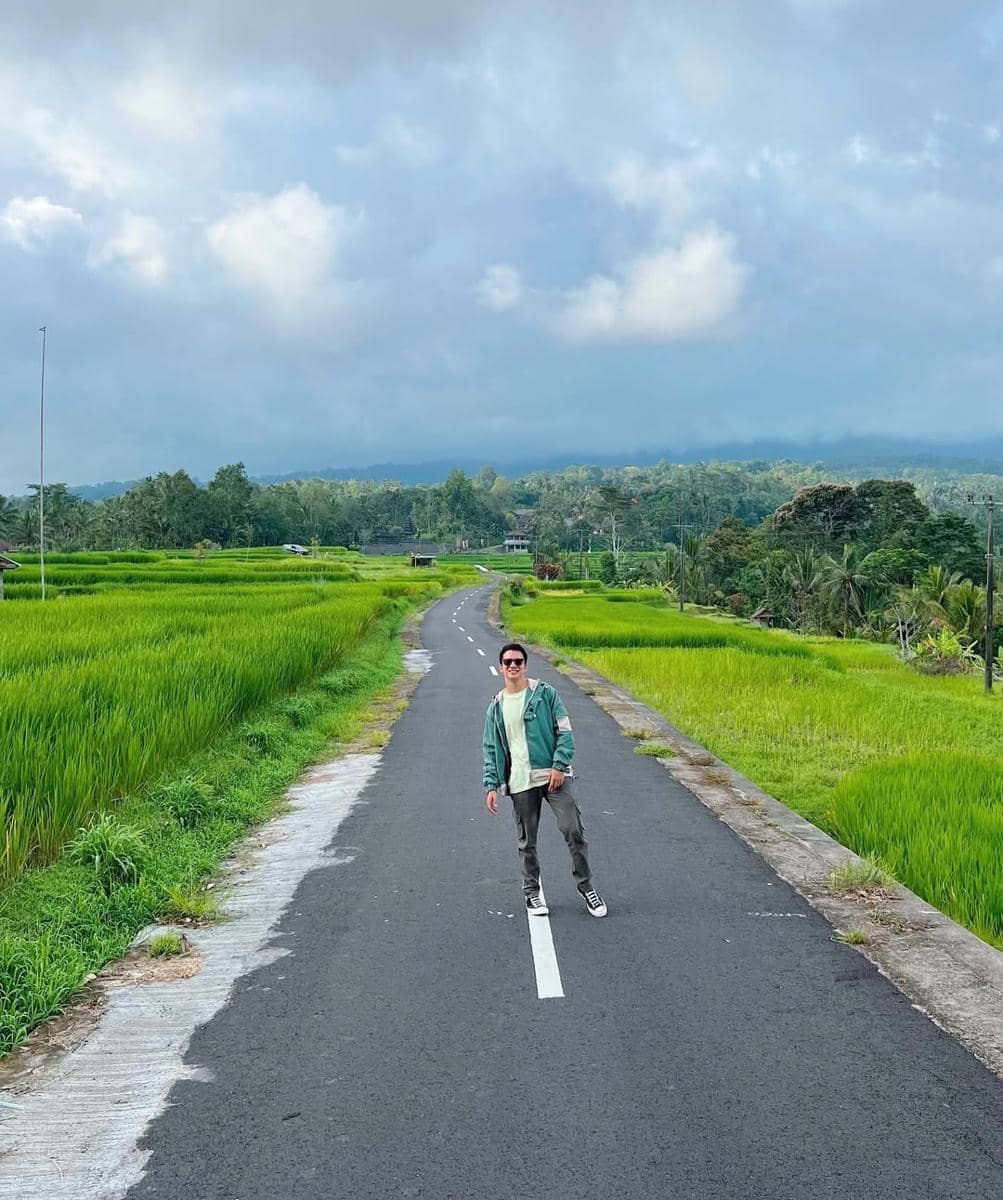 Seorang pria mengenakan jaket hijau dan kacamata berdiri di tengah jalan aspal yang membelah hamparan sawah hijau dengan latar langit berawan.