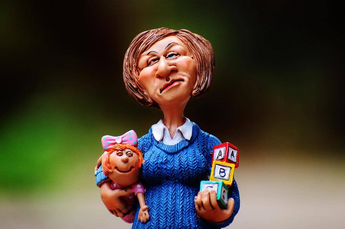 Figurine wanita mengenakan sweter biru memegang boneka bayi dan balok huruf warna-warni dengan latar belakang buram.