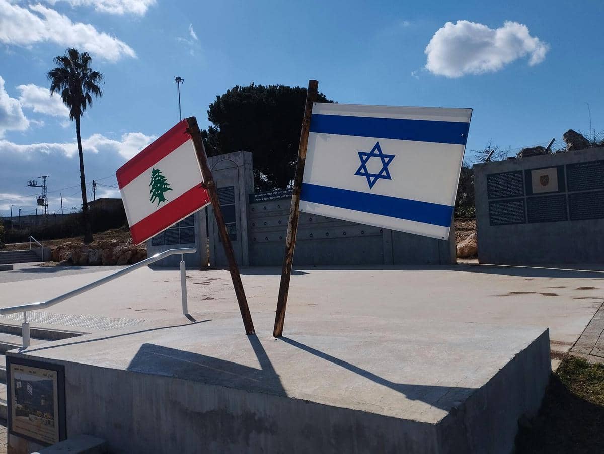 Bendera Lebanon dan Israel berdiri berdampingan di area terbuka dengan latar langit biru dan awan putih di siang hari.