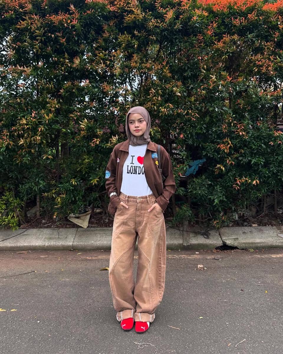 Seorang wanita berhijab mengenakan jaket cokelat, kaus putih bertuliskan I Love London, celana longgar krem, dan sneakers merah di luar ruangan.