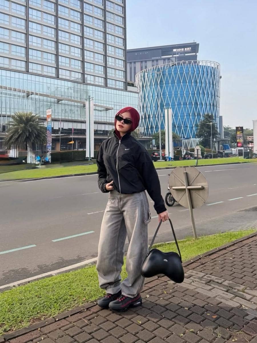 Seorang wanita bergaya kasual mengenakan jaket hitam, celana abu-abu longgar, dan sneakers hitam di area perkotaan dengan gedung tinggi di belakangnya.