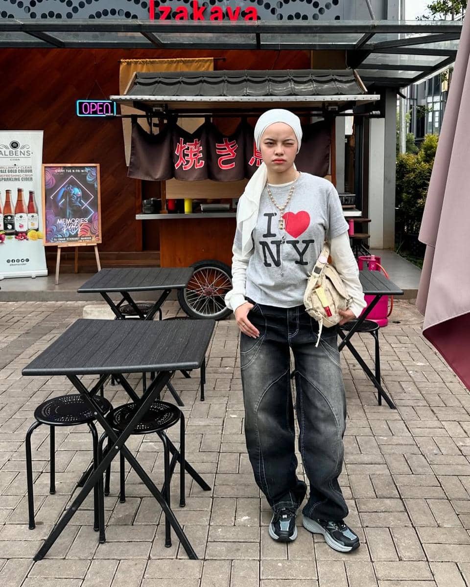 Seorang perempuan berhijab mengenakan kaus abu-abu bertulisan I Love NY, celana jeans longgar, dan sneakers hitam di depan kedai bergaya Jepang.