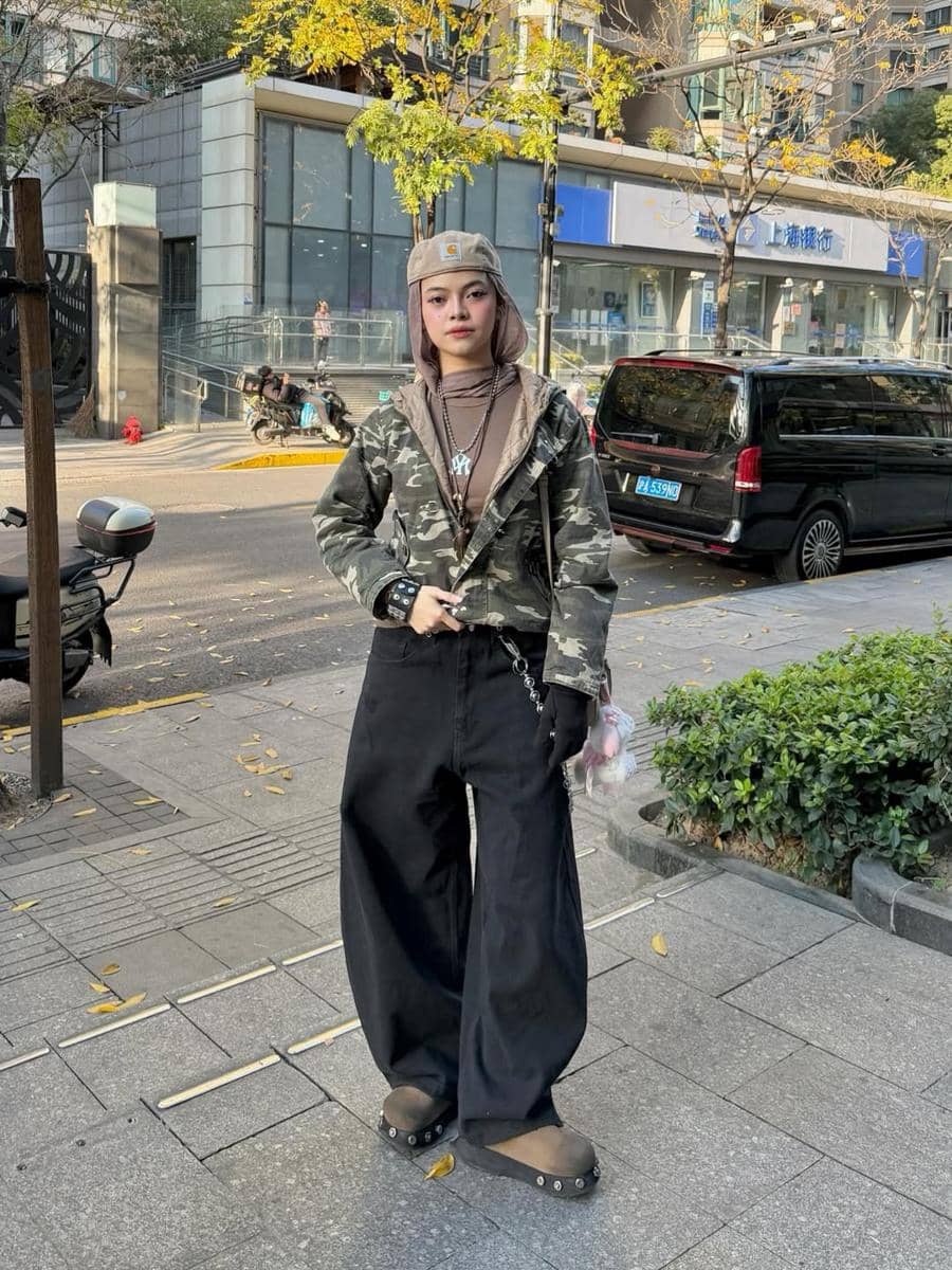 Seorang wanita bergaya street style mengenakan jaket motif kamuflase, celana hitam longgar, dan topi abu-abu di trotoar kota.