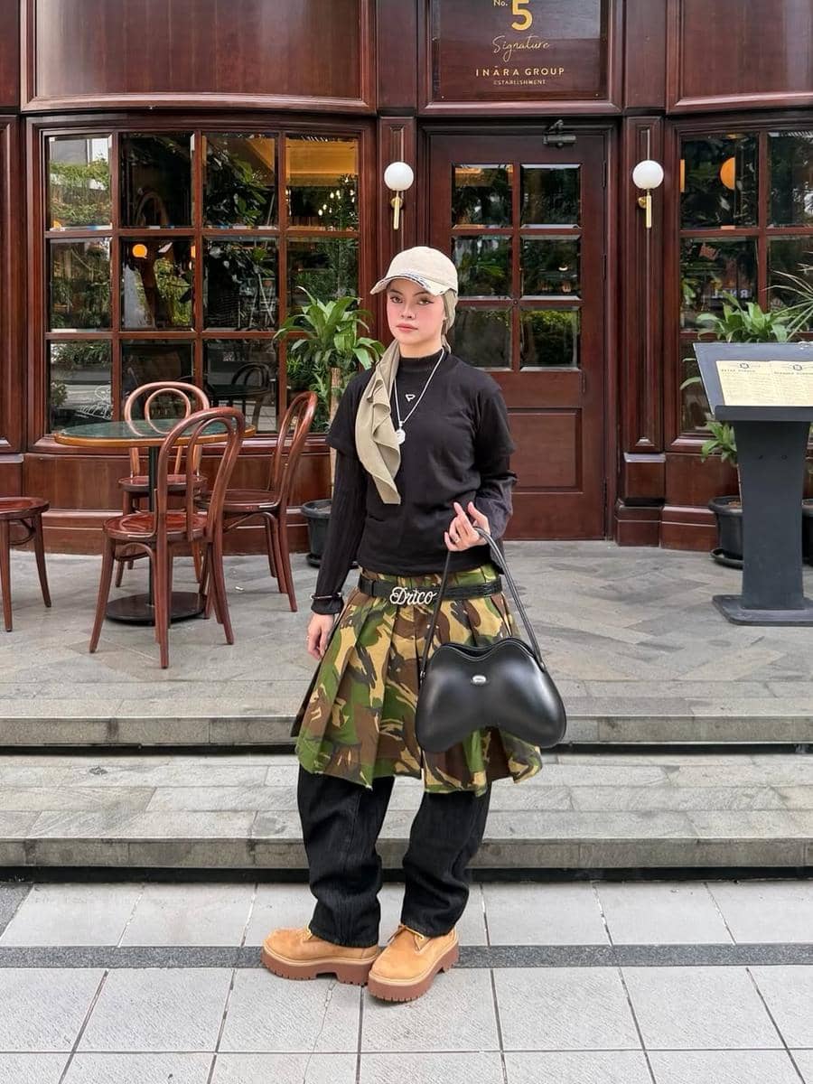 Seorang wanita berpose dengan gaya street style mengenakan topi krem, atasan hitam, rok motif kamuflase, celana longgar, dan sepatu cokelat di depan kafe bergaya klasik.