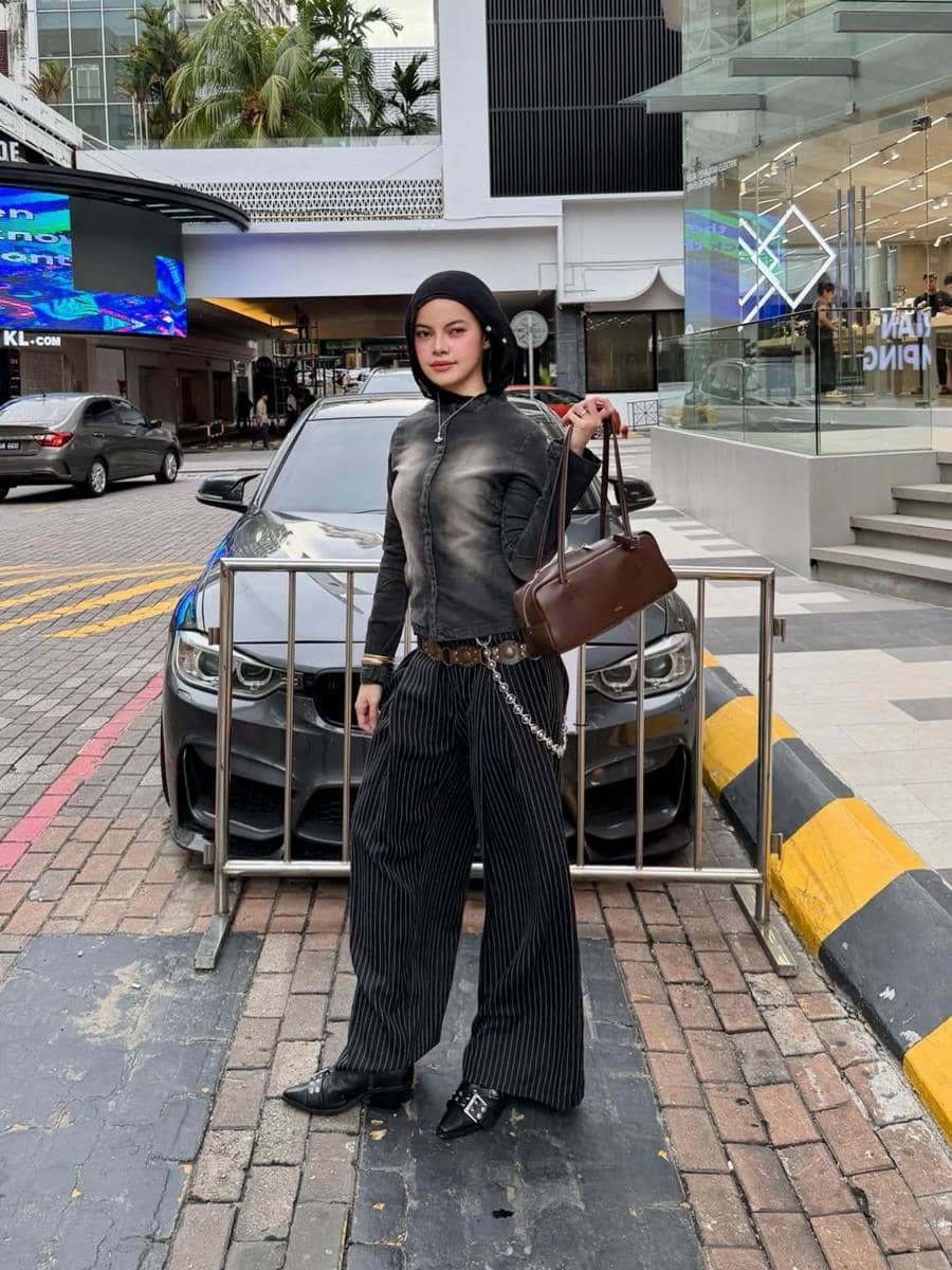 Seorang wanita berhijab bergaya street style mengenakan jaket hitam pudar, celana garis vertikal longgar, dan tas cokelat di area perkotaan.