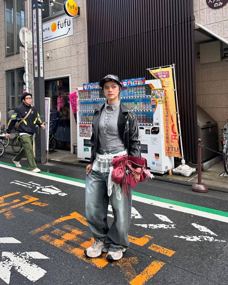 Seorang wanita bergaya street style mengenakan jaket kulit hitam, celana longgar, sneakers, dan topi di jalan kota dengan latar mesin penjual otomatis.