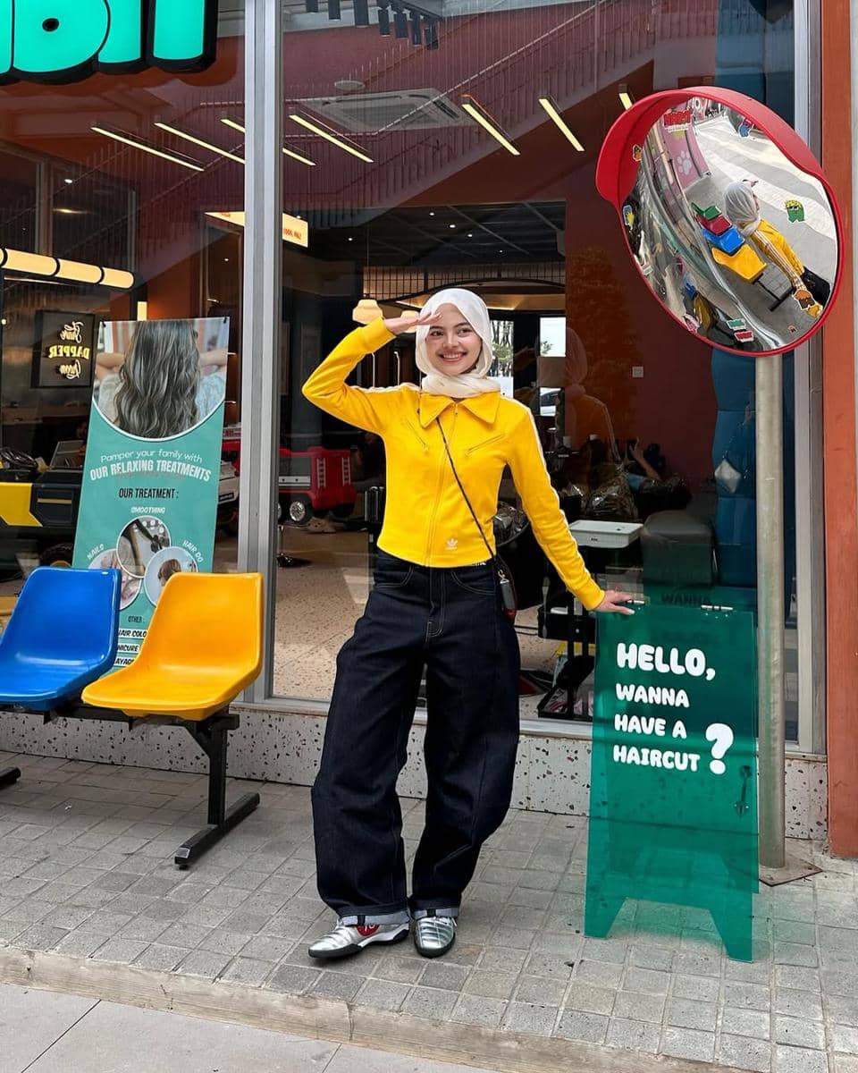 Seorang wanita berhijab mengenakan jaket kuning, celana jeans longgar, dan sneakers berpose di depan barbershop dengan papan hijau.