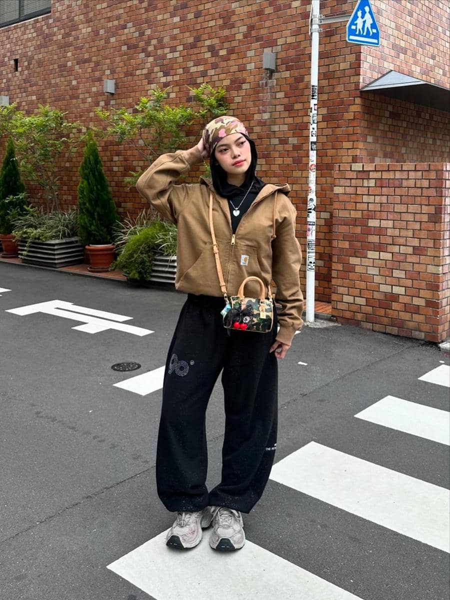 Seorang wanita bergaya kasual mengenakan hoodie cokelat, celana hitam longgar, sneakers abu-abu, dan tas kecil di jalan kota.