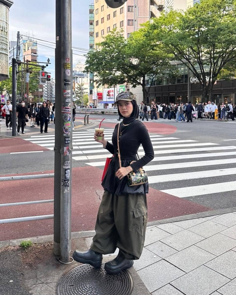 Seorang wanita bergaya street style mengenakan atasan hitam, celana kargo hijau longgar, dan topi di persimpangan jalan kota.