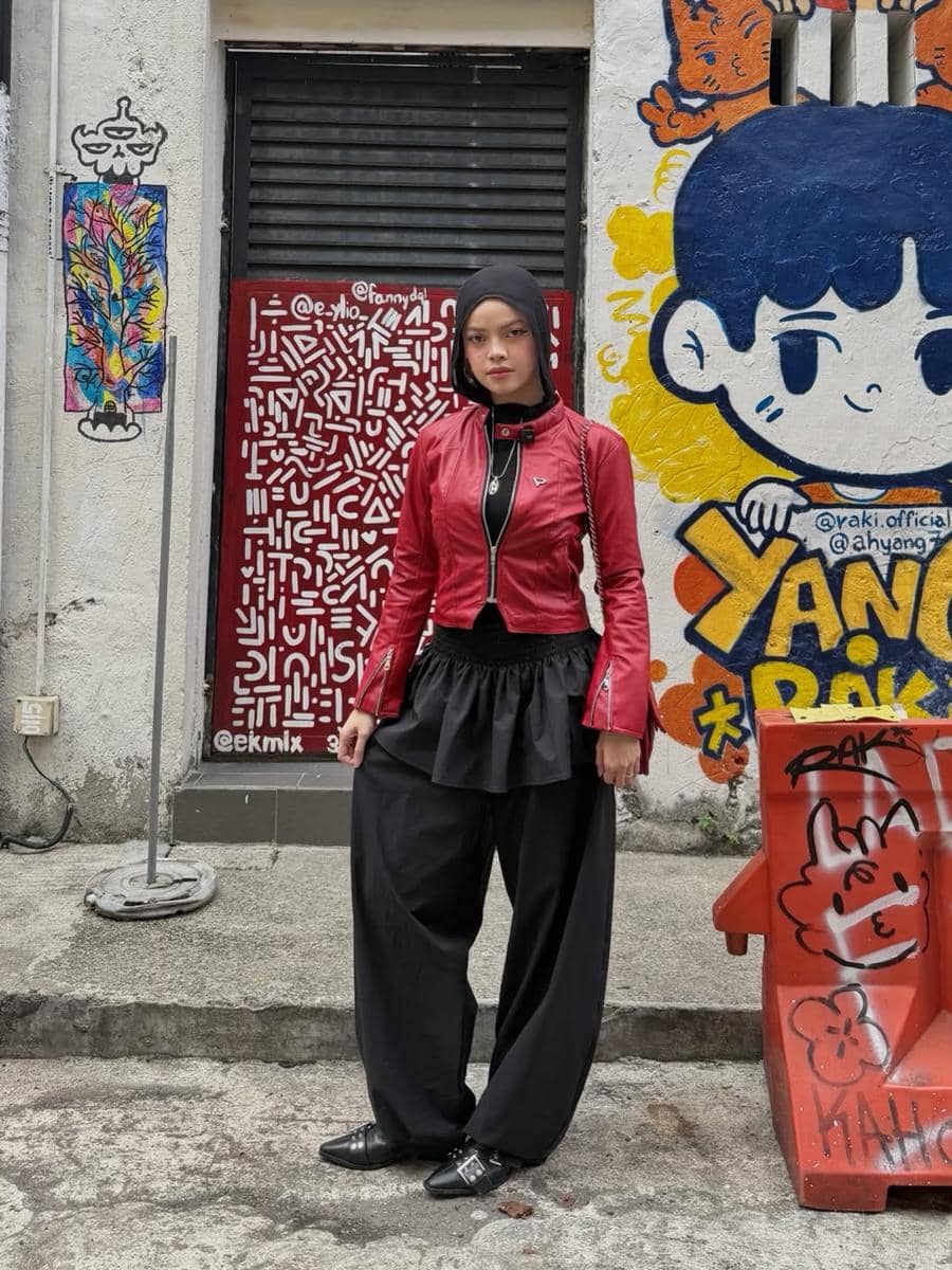 Seorang wanita berpose dengan gaya street style mengenakan jaket merah dan celana hitam longgar di depan dinding mural warna-warni.