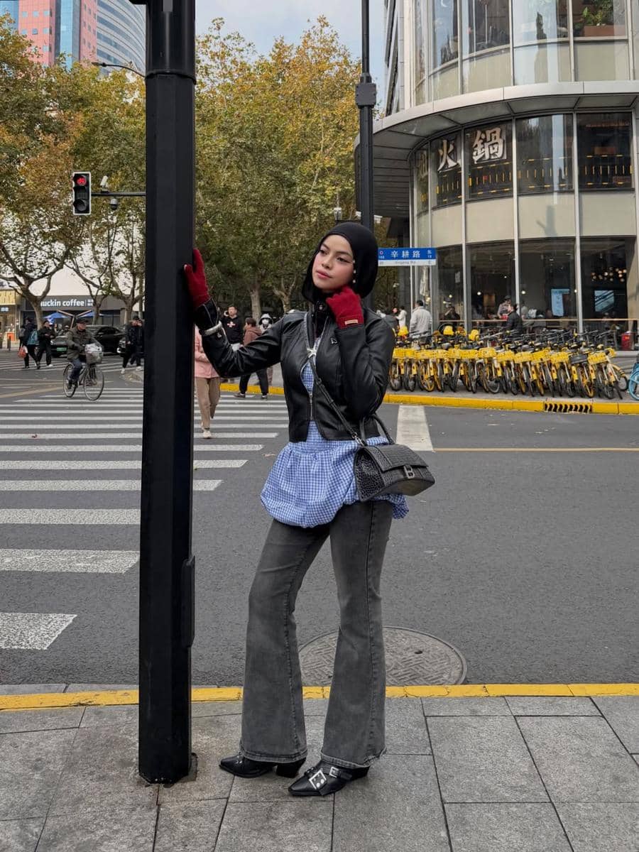 Seorang wanita berhijab bergaya street style mengenakan jaket kulit hitam, blus biru kotak, dan celana abu-abu di persimpangan jalan kota.