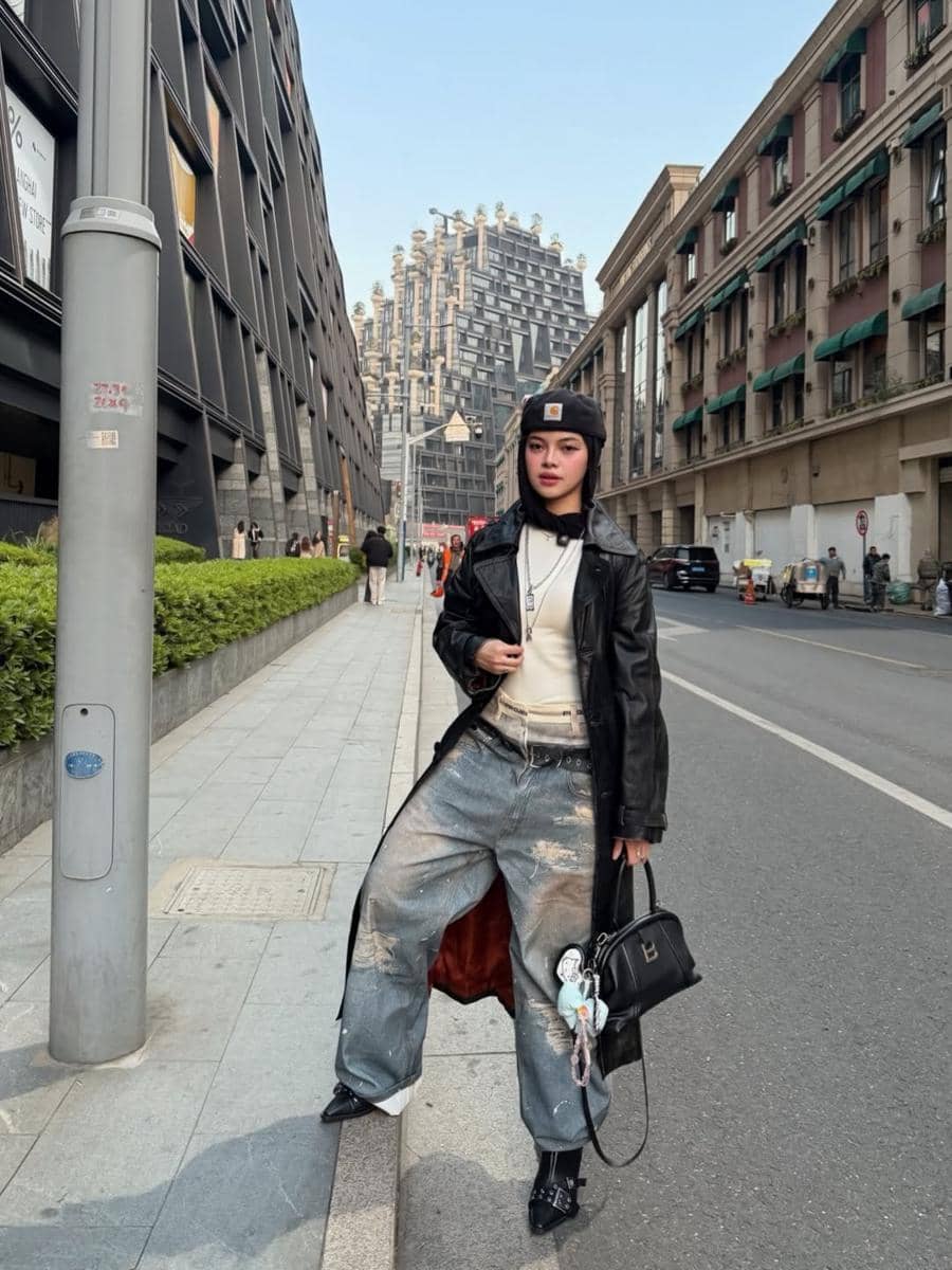 Seorang wanita bergaya street style mengenakan jaket kulit hitam panjang, celana jeans longgar, dan topi hitam di jalan perkotaan modern.