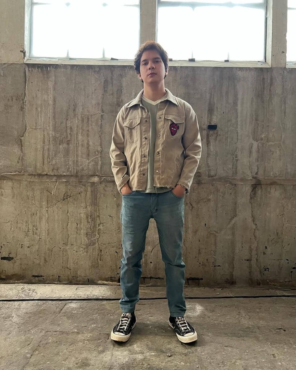 Seorang pria berdiri di depan dinding beton mengenakan jaket krem, kaus hijau muda, celana jeans biru, dan sepatu kets hitam putih.