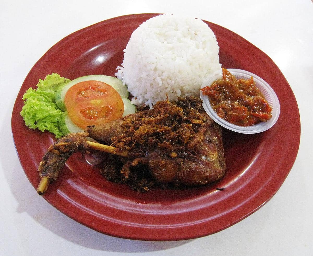 Sepiring bebek goreng disajikan dengan nasi putih, sambal, irisan tomat, mentimun, dan daun selada di atas piring merah.