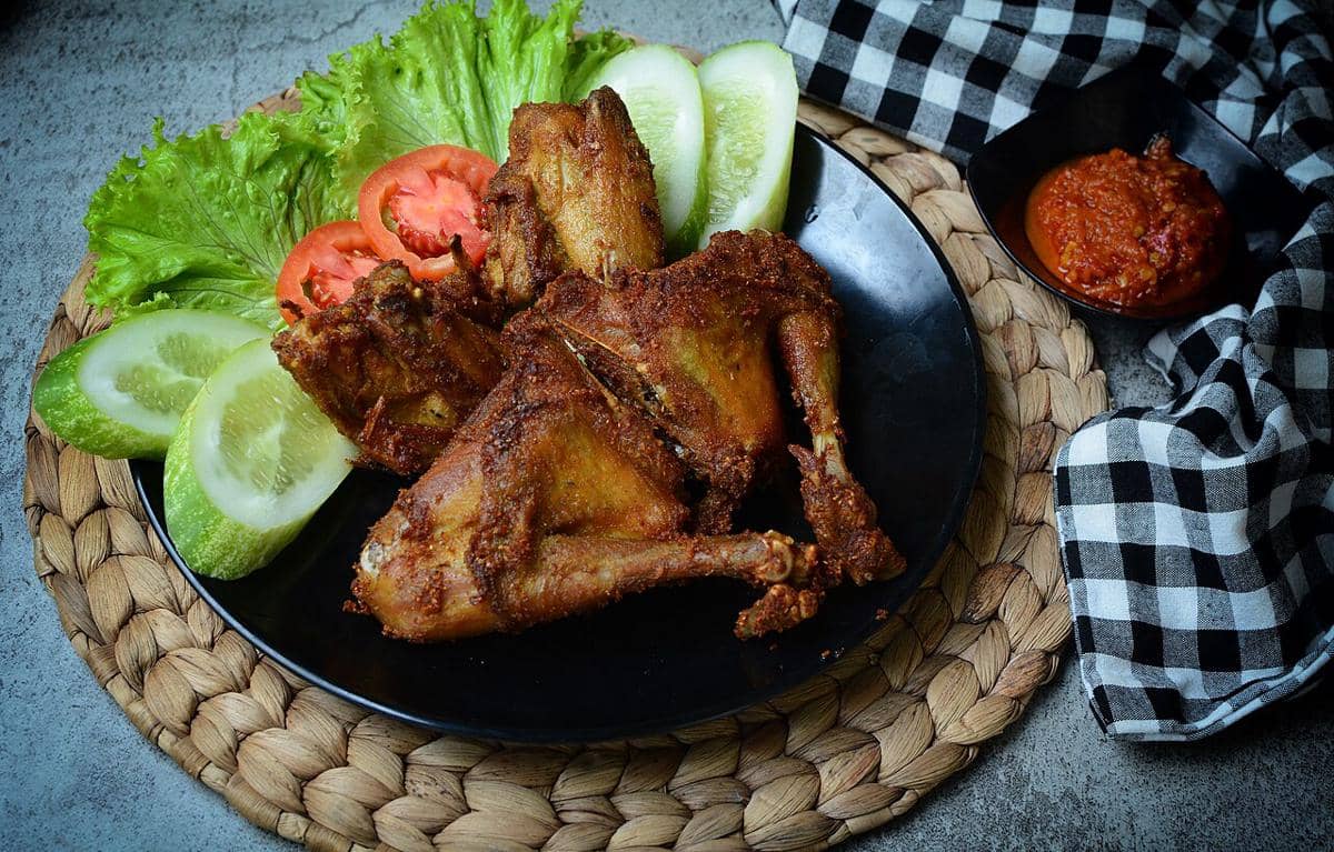 Ayam goreng berwarna keemasan disajikan di piring hitam dengan lalapan mentimun, tomat, daun selada, dan sambal di mangkuk kecil.