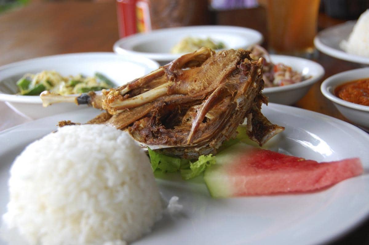 Sepiring bebek goreng disajikan dengan nasi putih, potongan semangka, dan lalapan di atas meja makan.