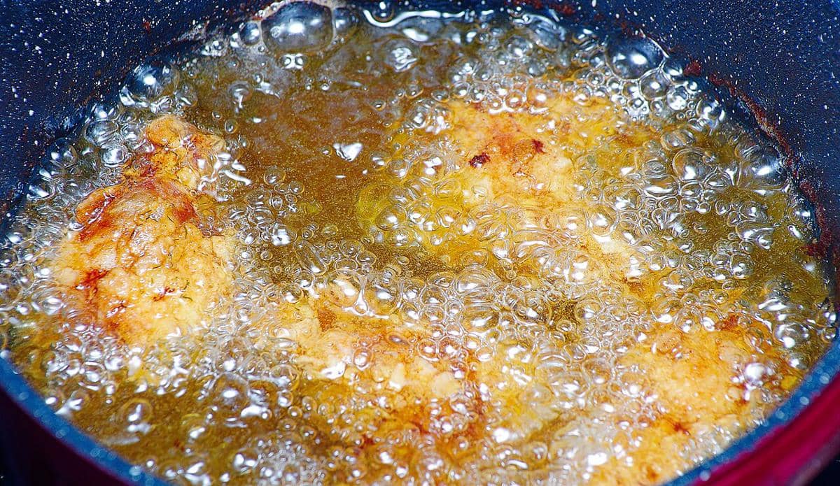 Potongan daging bebek sedang digoreng dalam minyak panas dengan gelembung minyak yang mendidih di wajan logam.