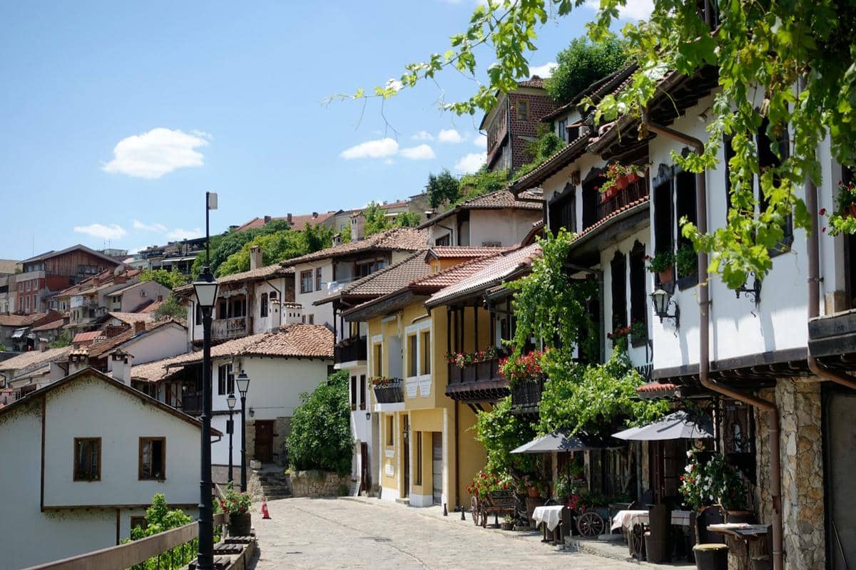 Deretan rumah tradisional dengan atap genteng merah di kawasan Kota Tua Veliko Tarnovo, dikelilingi tanaman hijau di bawah langit cerah.