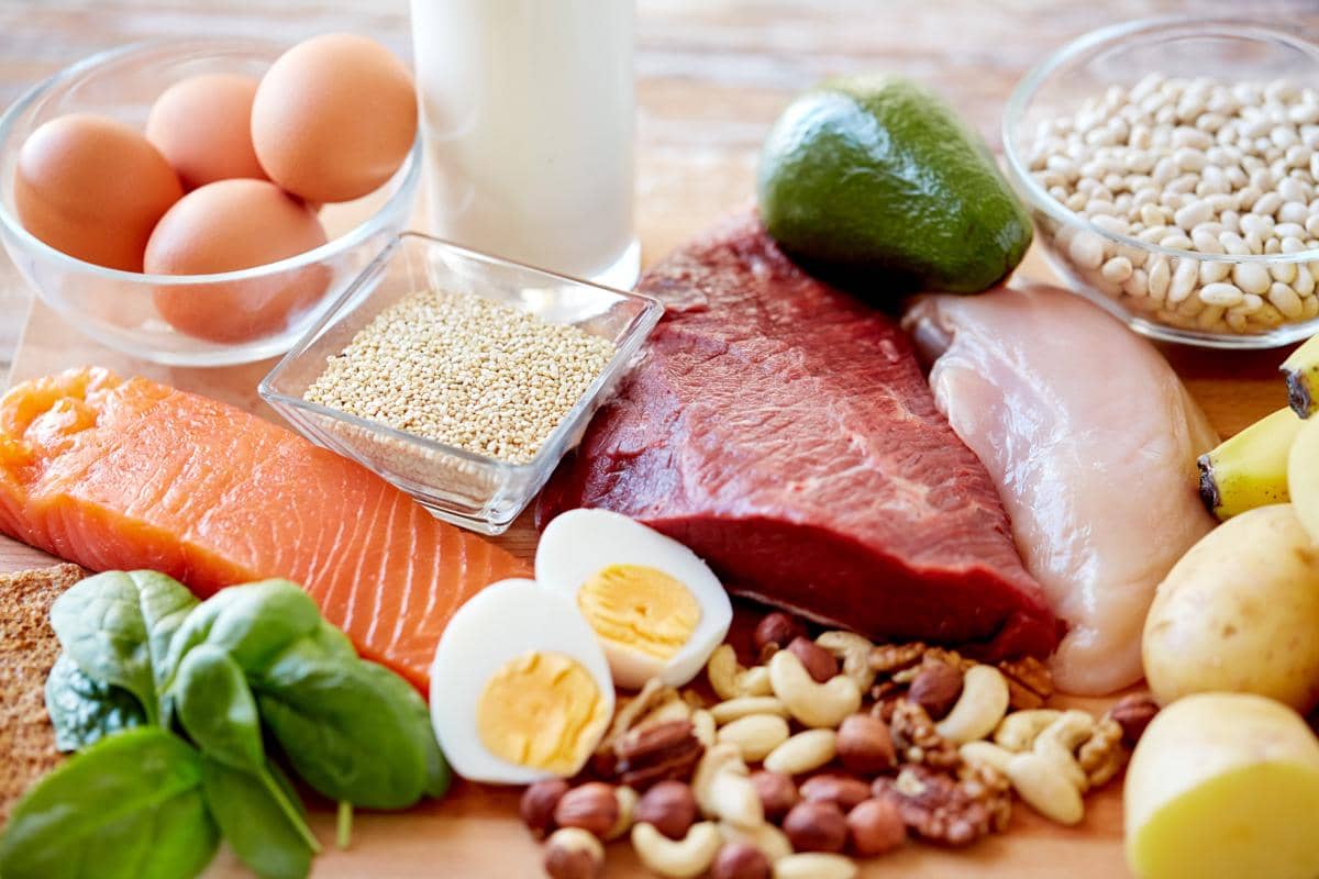 Beragam makanan sumber protein seperti telur, daging sapi, ayam, ikan salmon, kacang-kacangan, susu, dan sayuran hijau tersusun di atas meja kayu.
