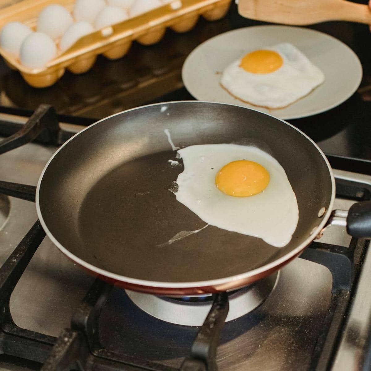 Telur sedang digoreng di atas wajan teflon di atas kompor gas dengan telur lain di piring dan wadah telur di latar belakang.