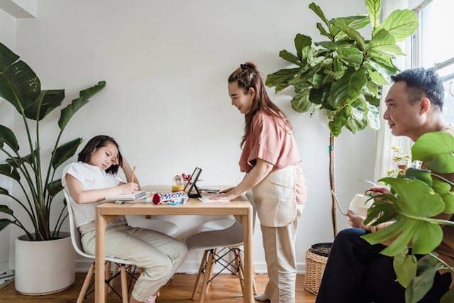 Tiga orang berada di ruang makan rumah dengan suasana santai, satu orang duduk di meja, satu berdiri melayani, dan satu lagi berbincang di dekat tanaman hias.