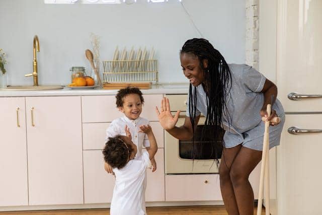 Seorang perempuan tersenyum sambil memberi tos kepada dua anak kecil di dapur modern yang terang dan rapi.