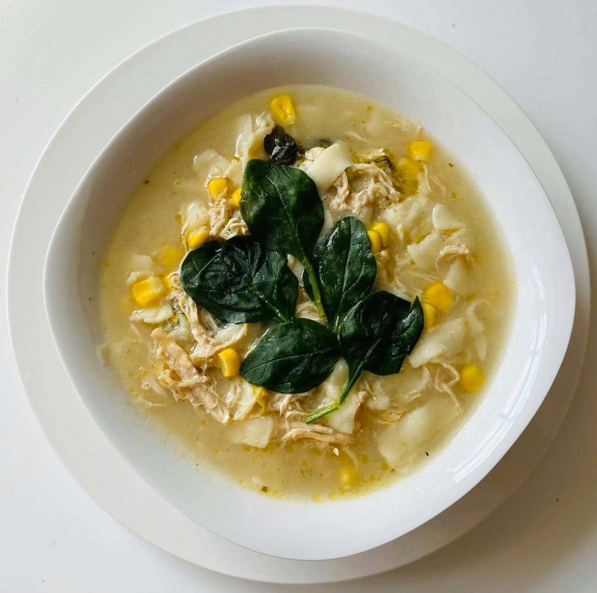 Semangkuk sup jagung hangat berisi jagung manis, suwiran ayam, potongan sayuran, dan daun bayam segar di atasnya.