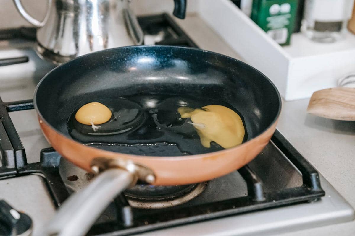 Wajan teflon berwarna tembaga di atas kompor gas dengan dua butir telur yang sedang dimasak di dapur modern.