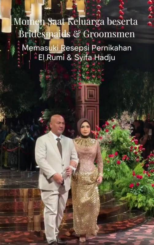 Seorang pria dan wanita berjalan beriringan di acara resepsi pernikahan dengan dekorasi bunga merah dan hijau yang mewah.