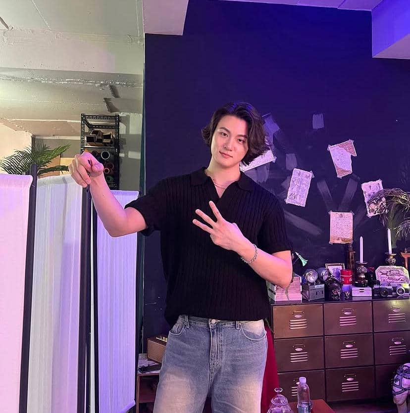 Seorang pria berambut sedang mengenakan kaus hitam dan jeans berpose sambil menunjukkan tiga jari di ruangan dengan pencahayaan ungu.