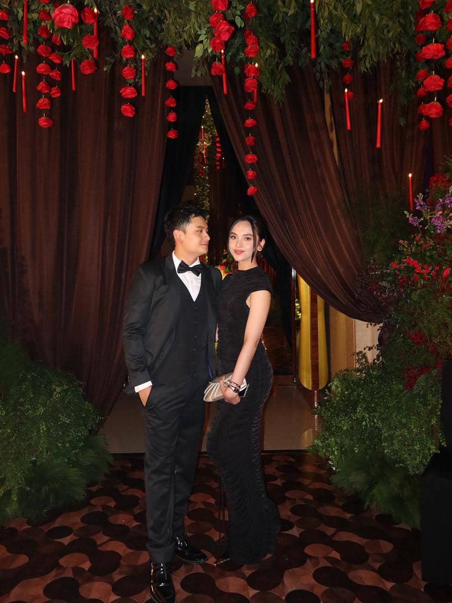 Sepasang tamu mengenakan busana formal hitam berpose di resepsi pernikahan dengan dekorasi bunga merah dan tirai cokelat elegan.